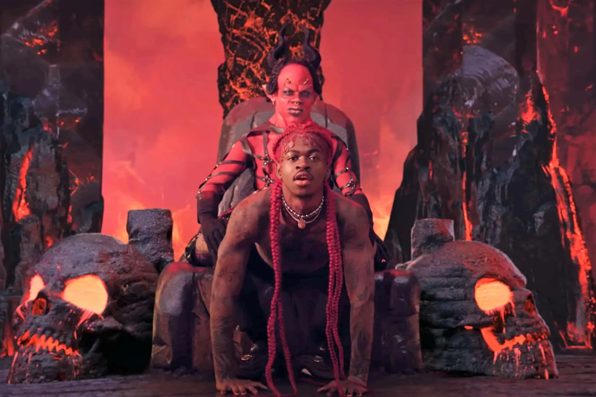 Image du clip Montero de Lil Nas X : le chanteur aux longues tresses rouges, à quatre pattes devant un trône de lave où siège un diable rouge à cornes, entre deux crânes géants aux yeux de feu.